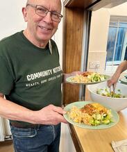 Volunteer takes two plates of food from the serving hatch while smiling